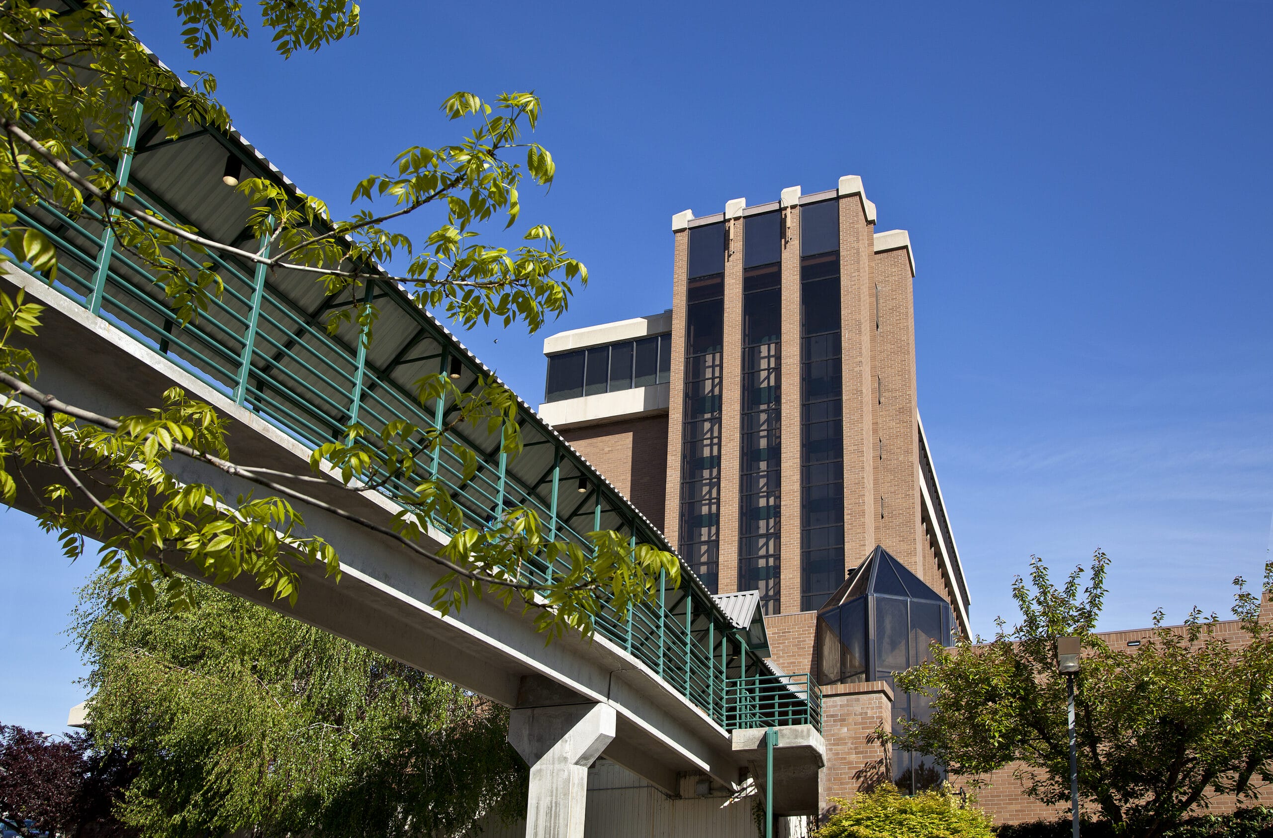 Pedestrian bridge next to brick building and trees.